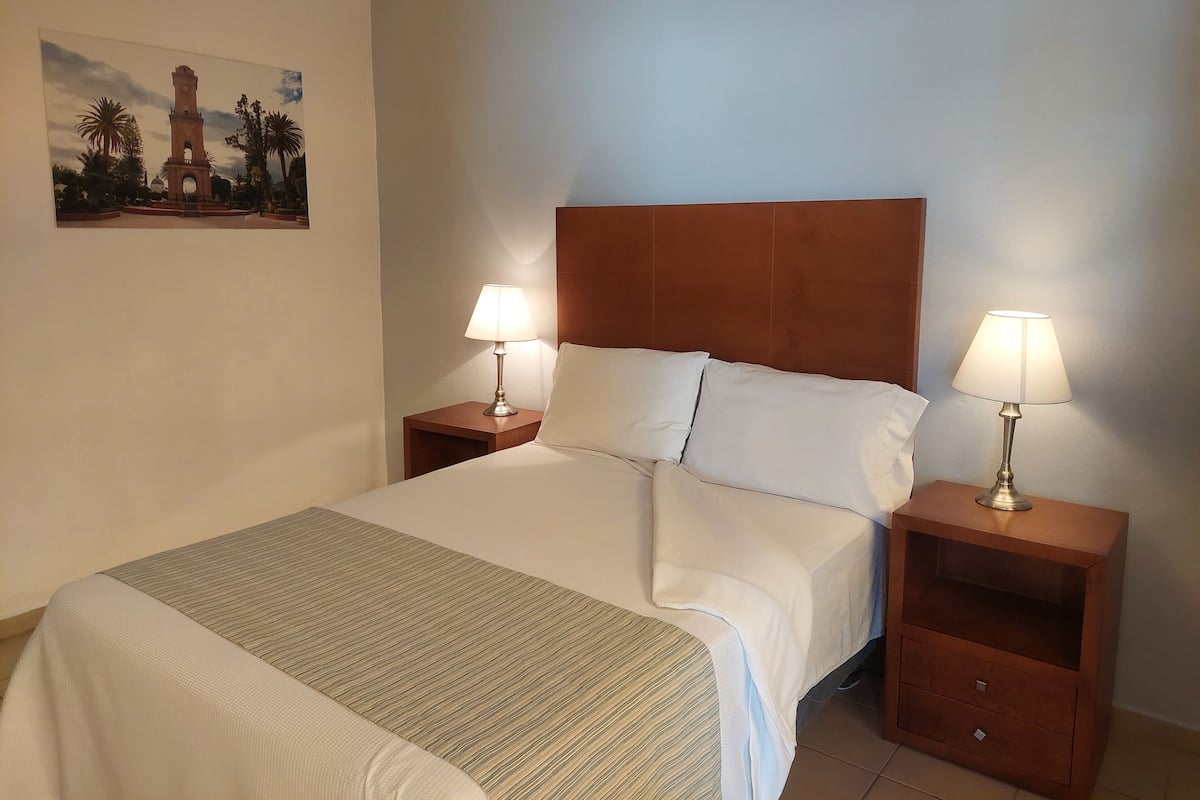 A hotel room with a large bed, two bedside tables with lamps, a headboard, and a painting on the wall.