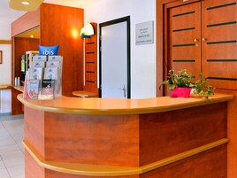 Un comptoir d’accueil d’hôtel avec un revêtement en bois, une enseigne bleue « ibis », un petit tabouret, une plante en pot et une porte avec une pancarte au-dessus.