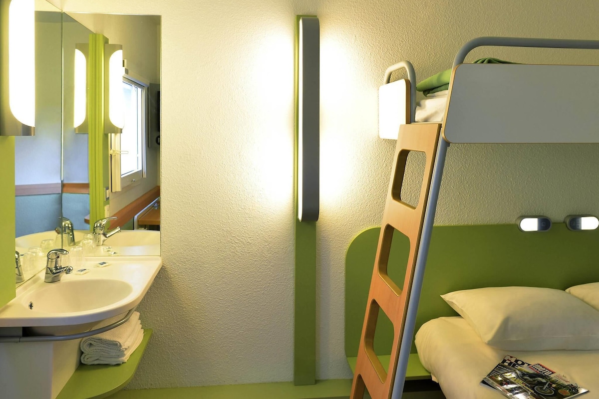 Habitación de hotel pequeña con literas, un lavamanos y un espejo, y una pared verde.