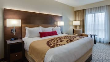 A hotel room with a large bed, two bedside lamps, a nightstand with a digital clock, a chair, a small table, and a large window with curtains.