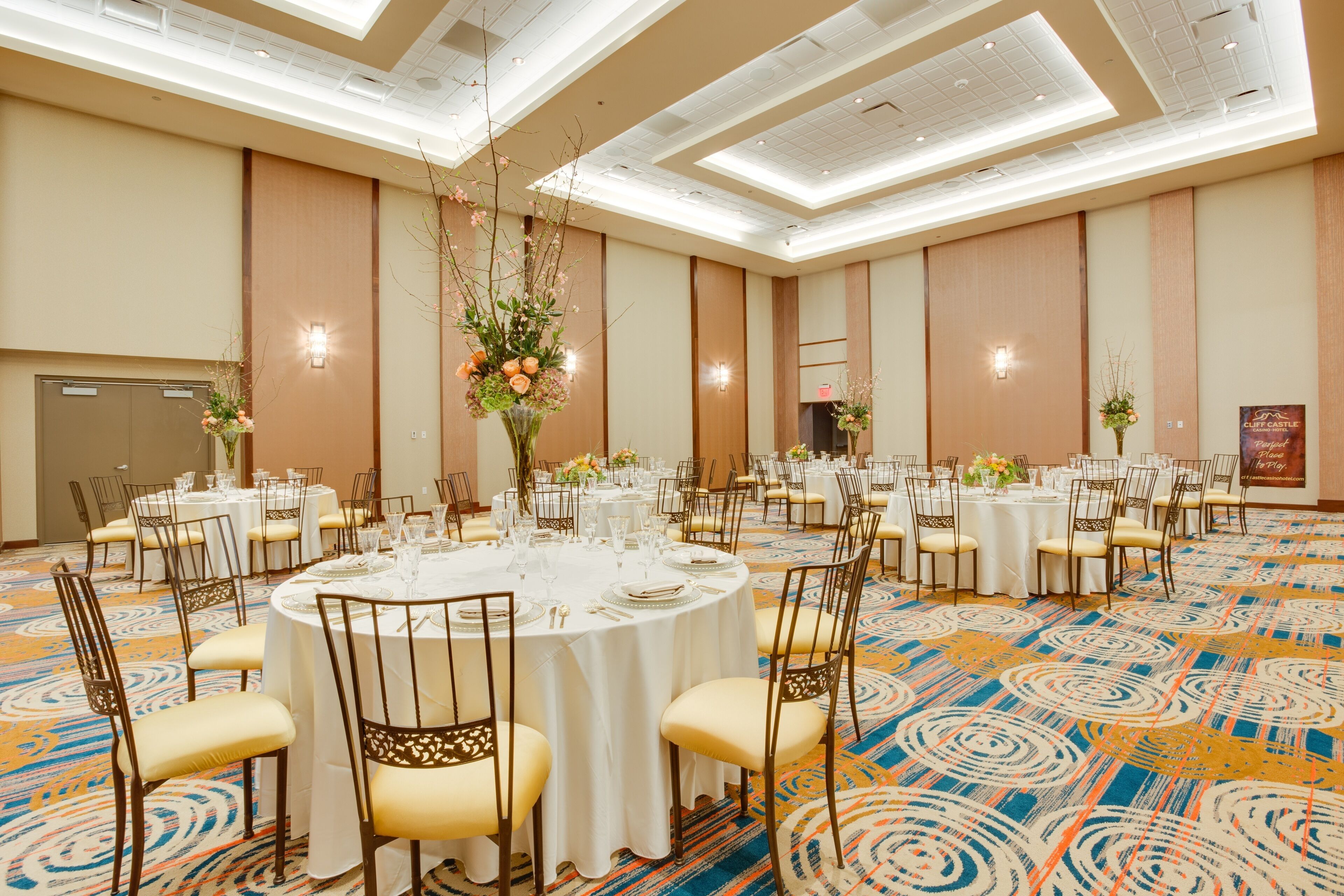 Een feestzaal met ronde tafels gedekt voor een diner, met witte tafelkleden en gouden stoelen.