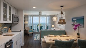 A modern kitchen with white cabinets and a built-in microwave. There is a dining area with a table set for two, a sofa, and a balcony with a view of the ocean.