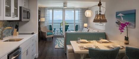 A modern kitchen with white cabinets and a built-in microwave. There is a dining area with a table set for two, a sofa, and a balcony with a view of the ocean.