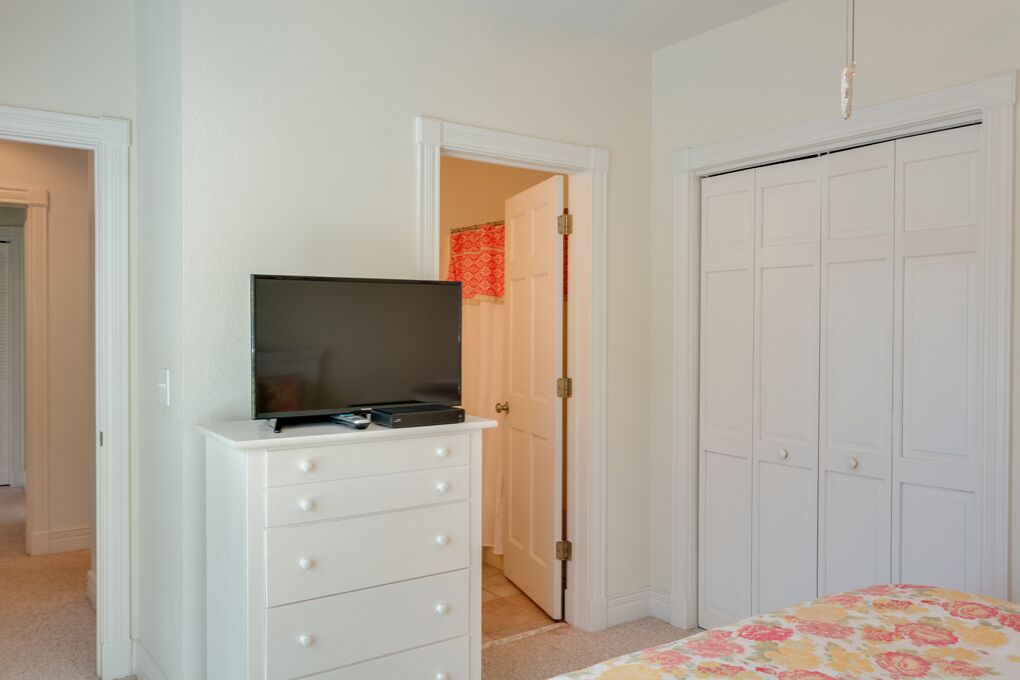 A bedroom with a white dresser, a television on top, a bed with a floral bedspread, and a white closet with a red door in the background.