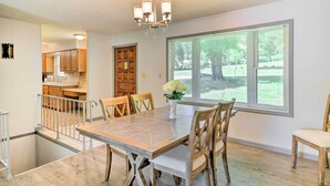 Un comedor con una mesa grande, sillas de madera y una araña. Se ve un jardín a través de un ventanal.