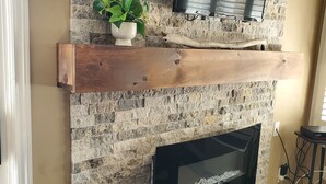 A stone fireplace with a TV mounted above it, a wooden shelf with a plant, and a dark upholstered chair in front.