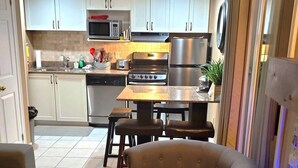 A compact kitchen with white cabinets, a microwave, a toaster, a stove, and a refrigerator. There is a dining table with four chairs and a sofa in the living area.