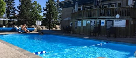Una piscina con bordo in legno, circondata da alberi e un edificio con posti a sedere all'aperto.