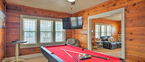A pool table with balls, a ceiling fan, and a television in a wooden-paneled room with windows.