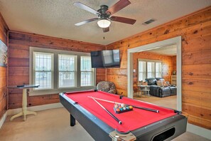 Une table de billard avec des billes, un ventilateur de plafond et un téléviseur dans une pièce aux murs lambrés et dotée de fenêtres.