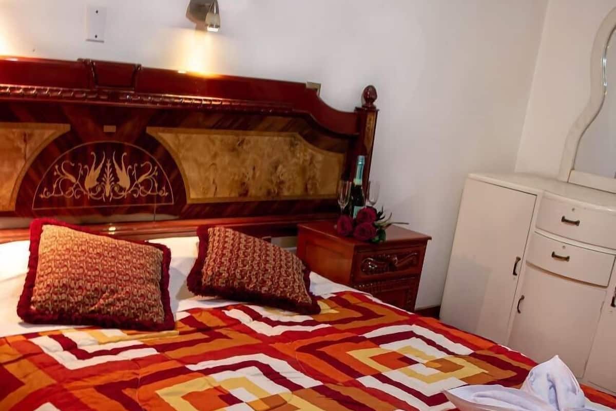 Un lit avec une couverture à motifs, des oreillers et une tête de lit ornée d’un motif décoratif. On y trouve une table de chevet avec une lampe, ainsi qu’une petite table sur laquelle est posé un vase de fleurs.
