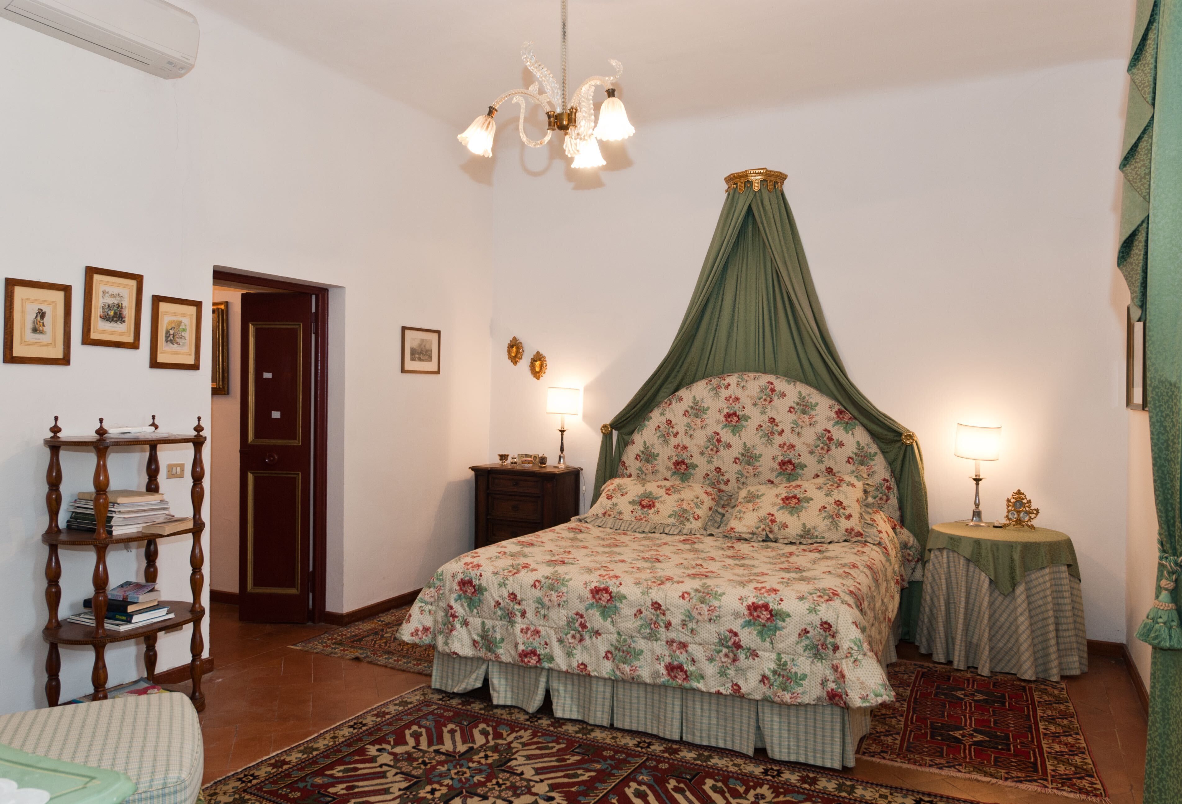 A bedroom with a floral bedspread, a canopy bed, a wooden side table, a nightstand, and a wall-mounted shelf with books.