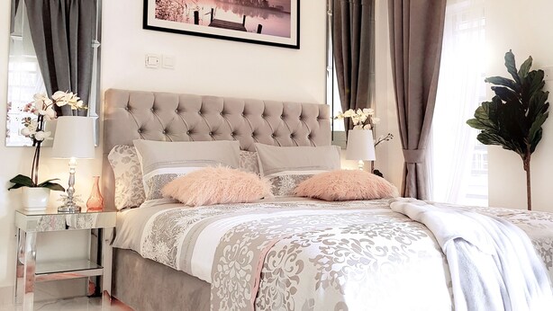 A bedroom with a tufted headboard, a large framed picture, a chandelier, and a large plant.