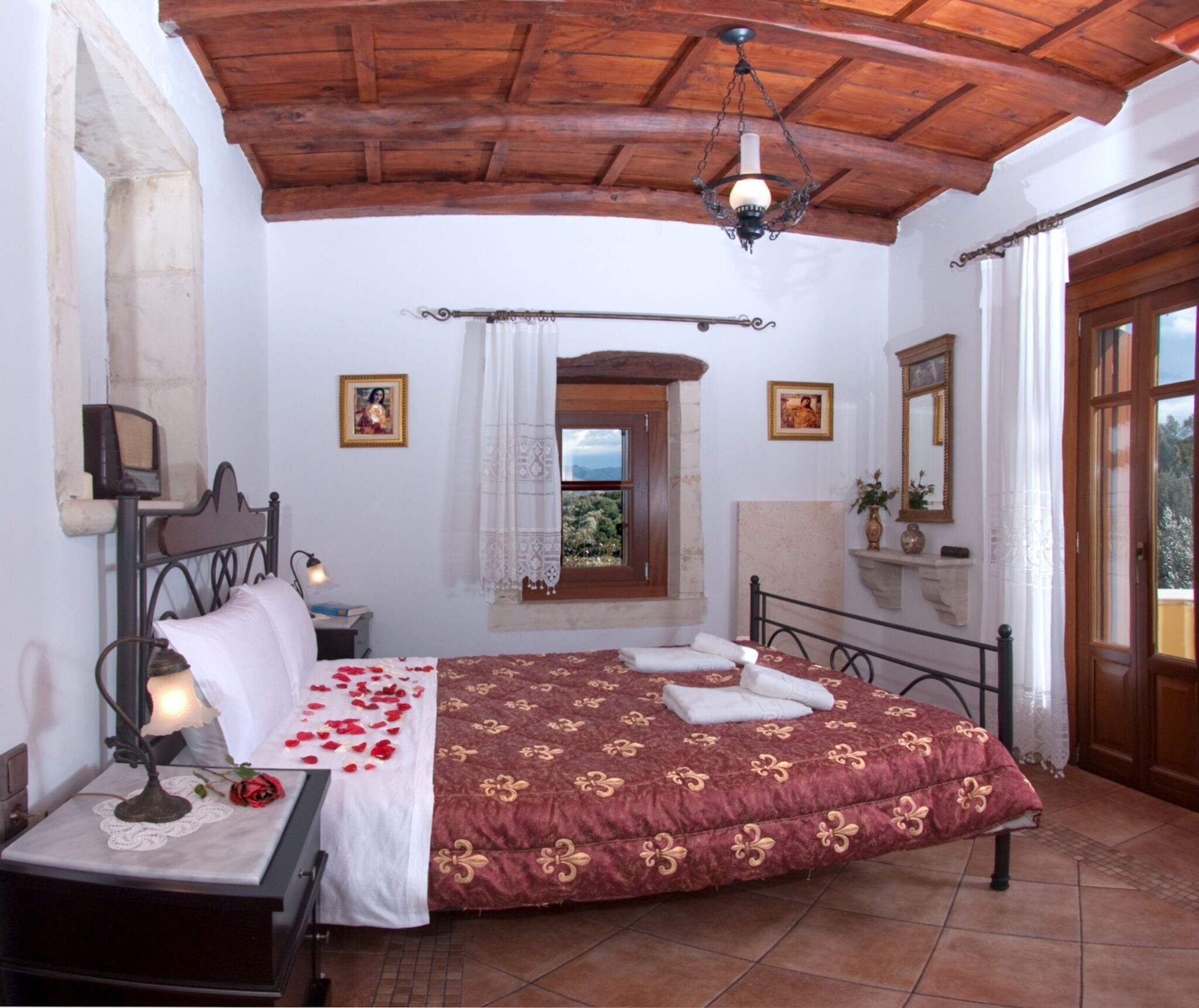 A bedroom with a wooden ceiling, a bed with a floral bedspread, a nightstand with a lamp, and a window with a view of greenery.