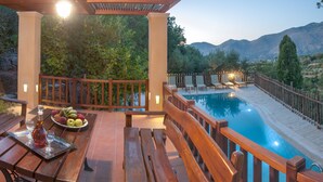 A wooden deck with a pool, a table with fruit and a bottle, and a view of mountains. - Private villa with private pool and amazing view in an unspoilt environment (Rethymno)