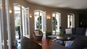 A spacious living room with a grey sofa, a wicker chair, and a coffee table. There are multiple open doors leading to a garden.