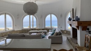 Ett rymligt vardagsrum med en stor säng, två soffor och en öppen spis. Det finns tre välvda fönster som ger utsikt över havet.