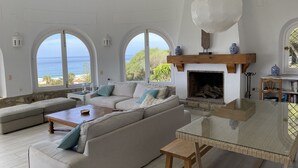 Ett rymligt vardagsrum med en stor soffa, ett soffbord i glas och en öppen spis. Det finns tre stora fönster som erbjuder utsikt över havet.