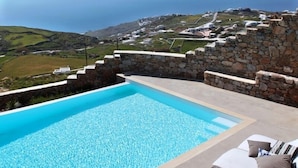 A swimming pool with lounge chairs and a stone wall, overlooking a coastal landscape. - Villa DeLucien (Mykonos)