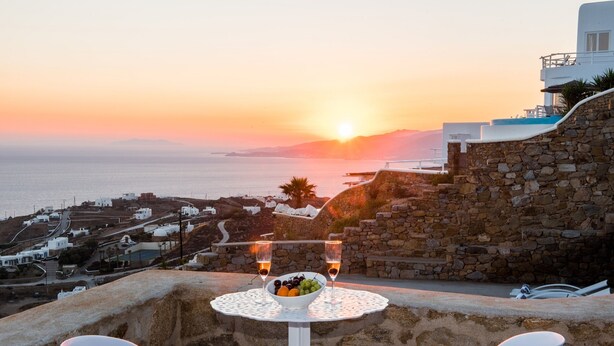 A table with a bowl of fruit and two glasses of wine, overlooking a coastal town at sunset. - Villa DeLucien (Mykonos)