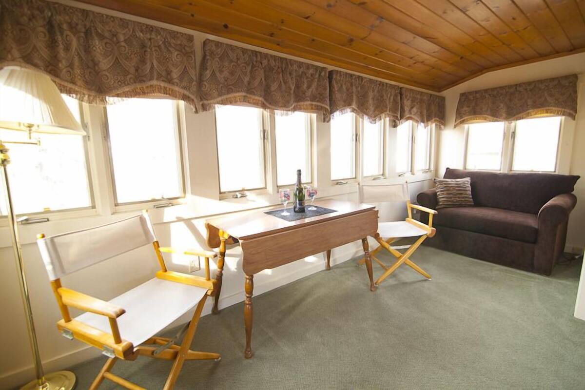 Ein Zimmer mit Holzboden, einem Schreibtisch mit Lampe, einem braunen Sofa und einem Fenster mit Vorhängen.