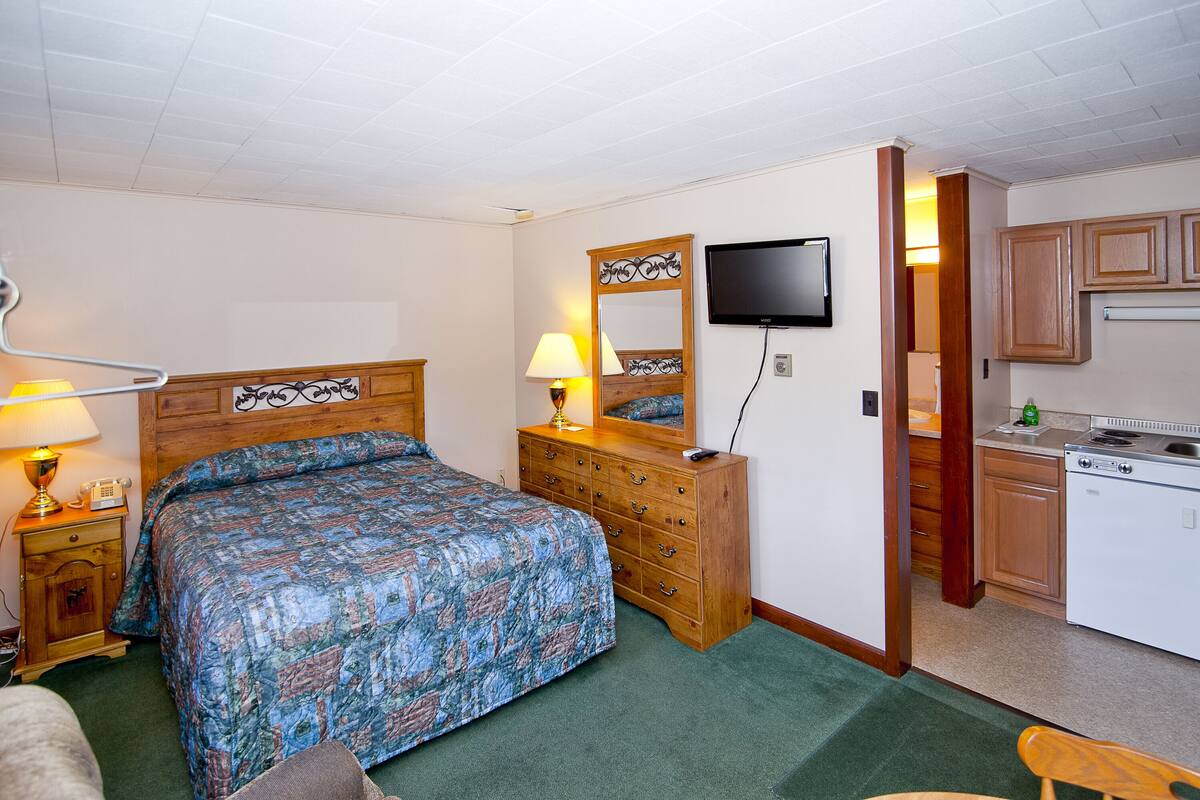 Ein Schlafzimmer mit Bett, Kommode, Spiegel, Fernseher und einer Küchenzeile.