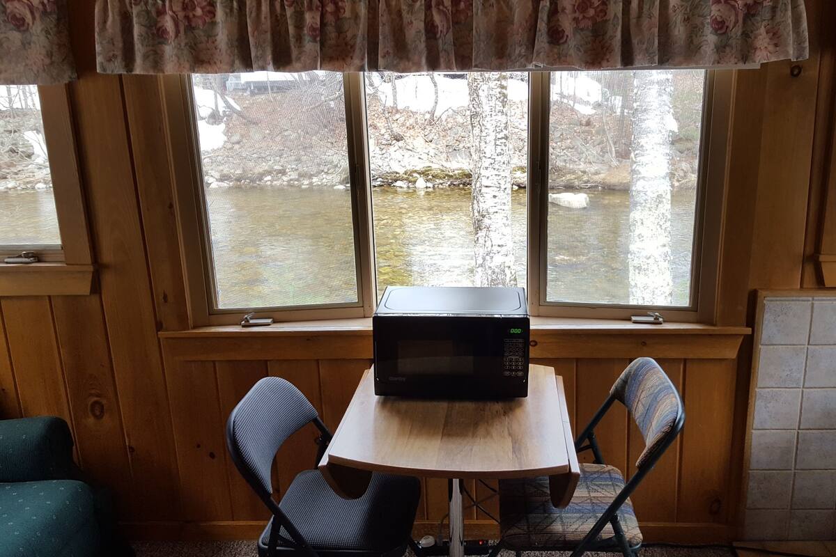 Ein Holzinterieur mit Tisch und Stühlen, einer Mikrowelle auf dem Tisch und Blick auf eine felsige Landschaft durch große Fenster.
