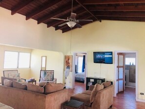 Un salón amplio con ventilador de techo, un conjunto de sofás, un televisor montado en la pared y una puerta que da a otra habitación.