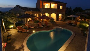 A swimming pool with lounge chairs and umbrellas, surrounded by a stone building with warm lighting.