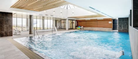 An indoor swimming pool with a wooden ceiling, clear glass walls, and a view of the outside.