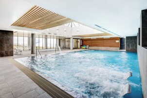 Una piscina interior con techo de madera, paredes de vidrio transparente y vistas al exterior.