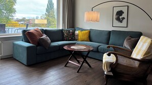 A modern living room with a sectional sofa, a round coffee table, and a wicker chair. A large window offers a view of the cityscape.