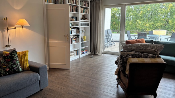 A living room with a grey sofa, a patterned armchair, a bookshelf, and a view of the outdoors through large windows.