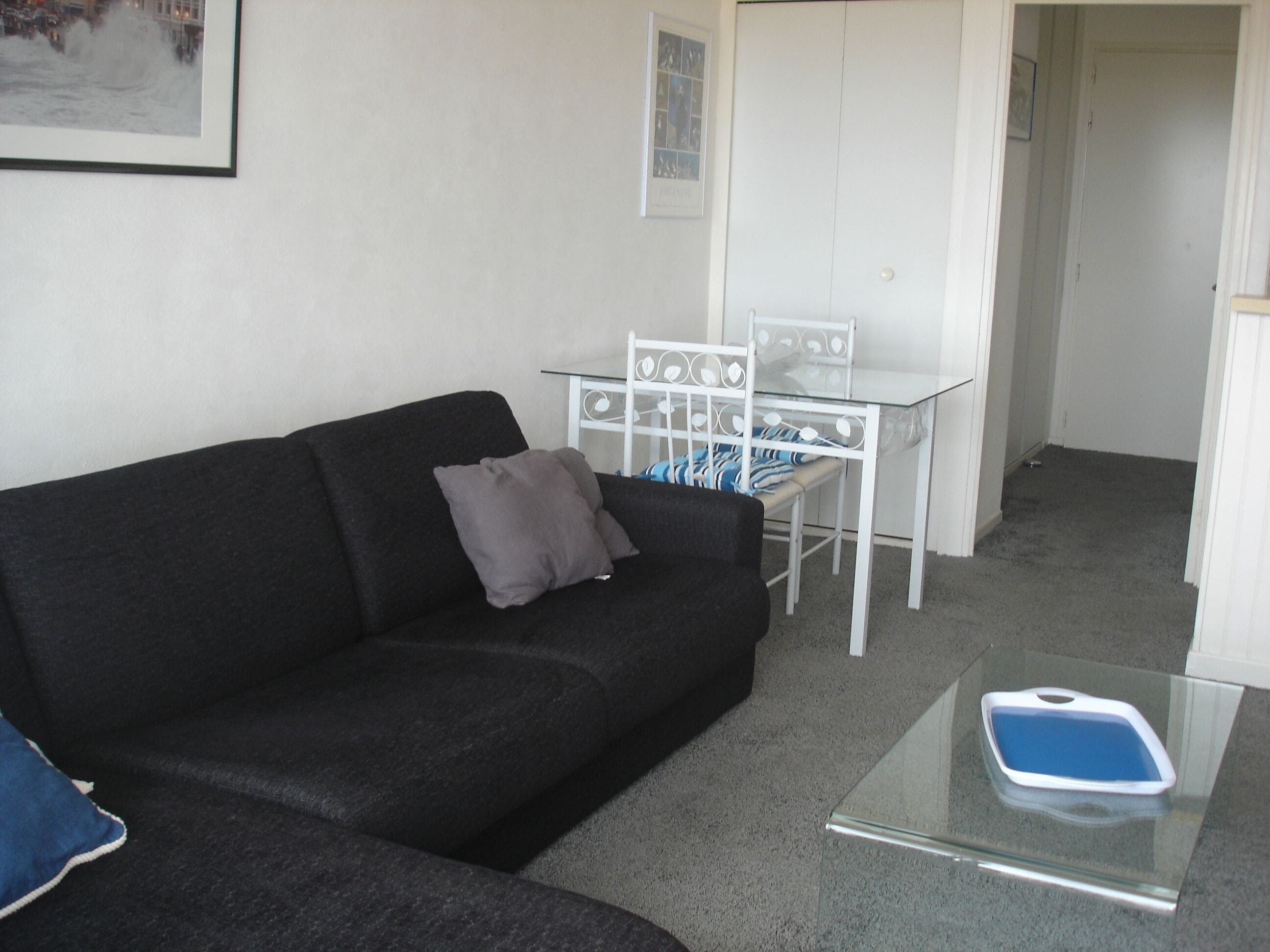 A living room with a black sofa, a glass coffee table, a white metal table with chairs, and a framed picture on the wall.