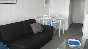 Ein Wohnzimmer mit einem schwarzen Sofa, einem Glastisch, einem weißen Metalltisch mit Stühlen und einem gerahmten Bild an der Wand.