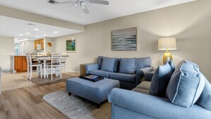 A living room with a sofa, ottoman, and a lamp. There is a dining area with a table and chairs. - Surf Court 30 - Updated 2 Bedroom Condo - Short Walk to Beach (Hilton Head Island)