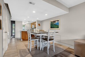A dining area with a white table and chairs, a kitchen in the background, and a framed picture on the wall.