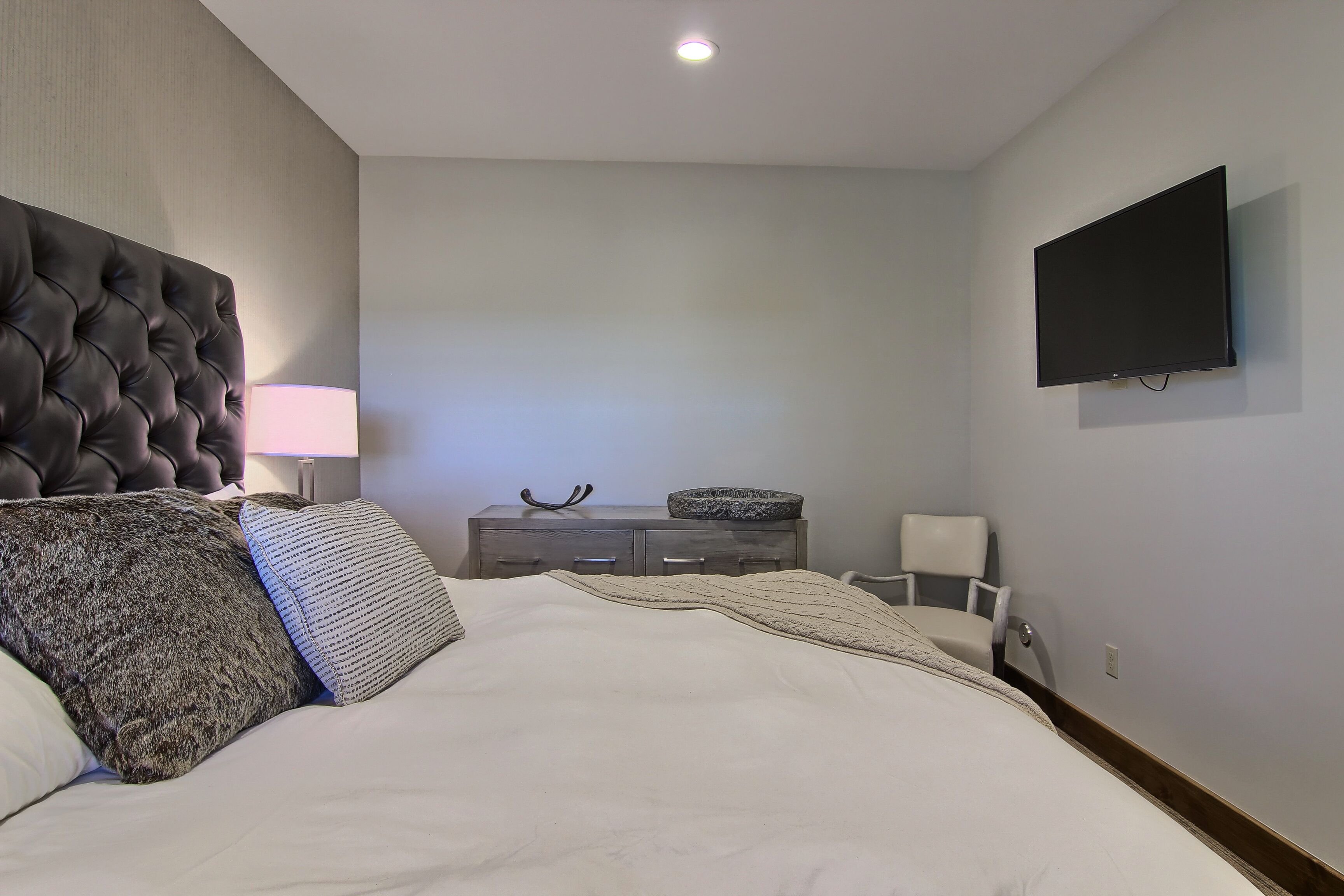 A bedroom with a large bed, a headboard, a bedside table with a lamp, a dresser, a television mounted on the wall, and a chair.