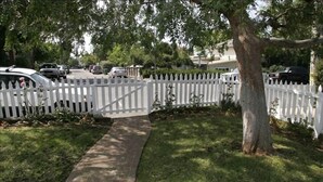 A residential street with a white picket fence, a brick pathway, and parked cars.