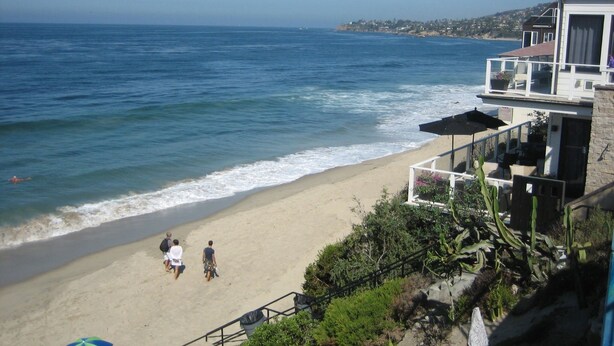 A beach with a view of the ocean, a building with a balcony overlooking the beach, and a few people walking along the shore.