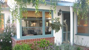 A house with a brick foundation, a window with a view of the interior, and a garden with various plants and flowers.