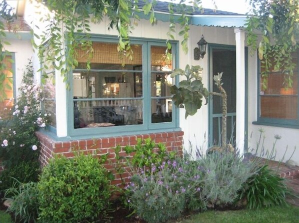 A house with a brick foundation, a window with a view of the interior, and a garden with various plants and flowers.