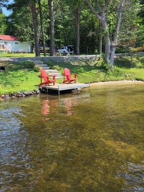 A dock with two red Adirondack chairs, a clear pond, and a white vehicle parked in the background.