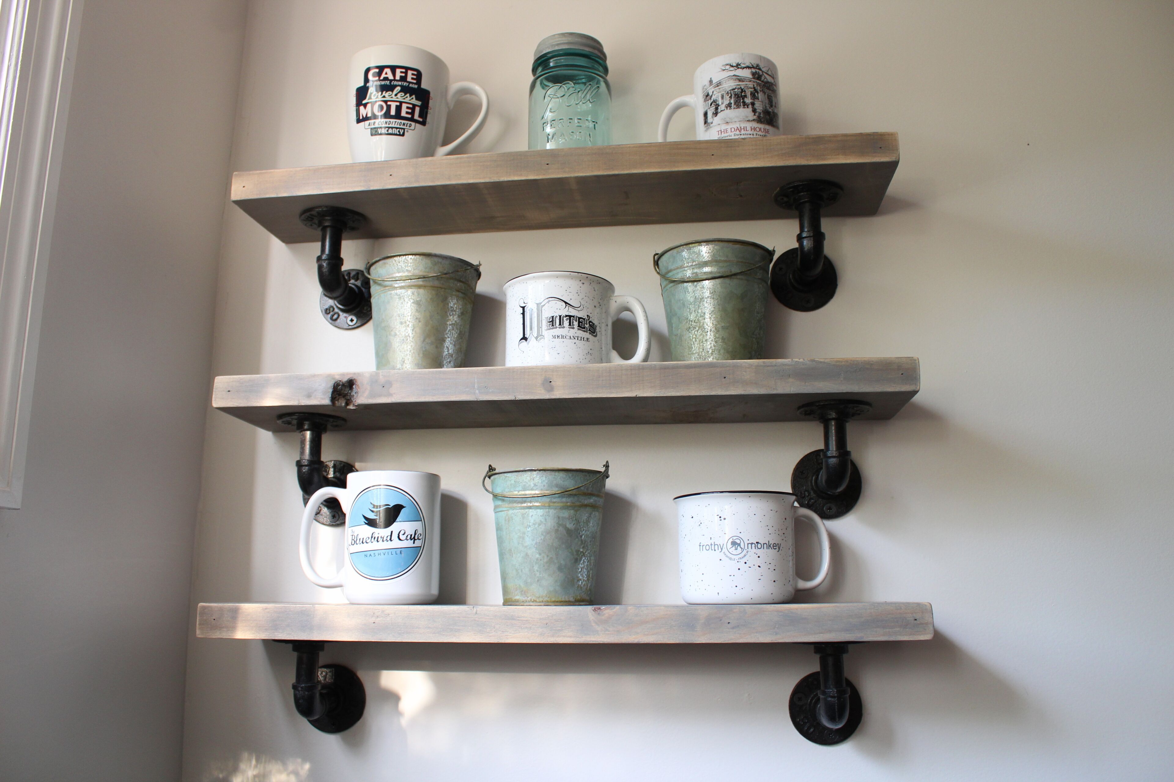 Three wooden shelves mounted on a wall, each holding various mugs and a metal bucket.