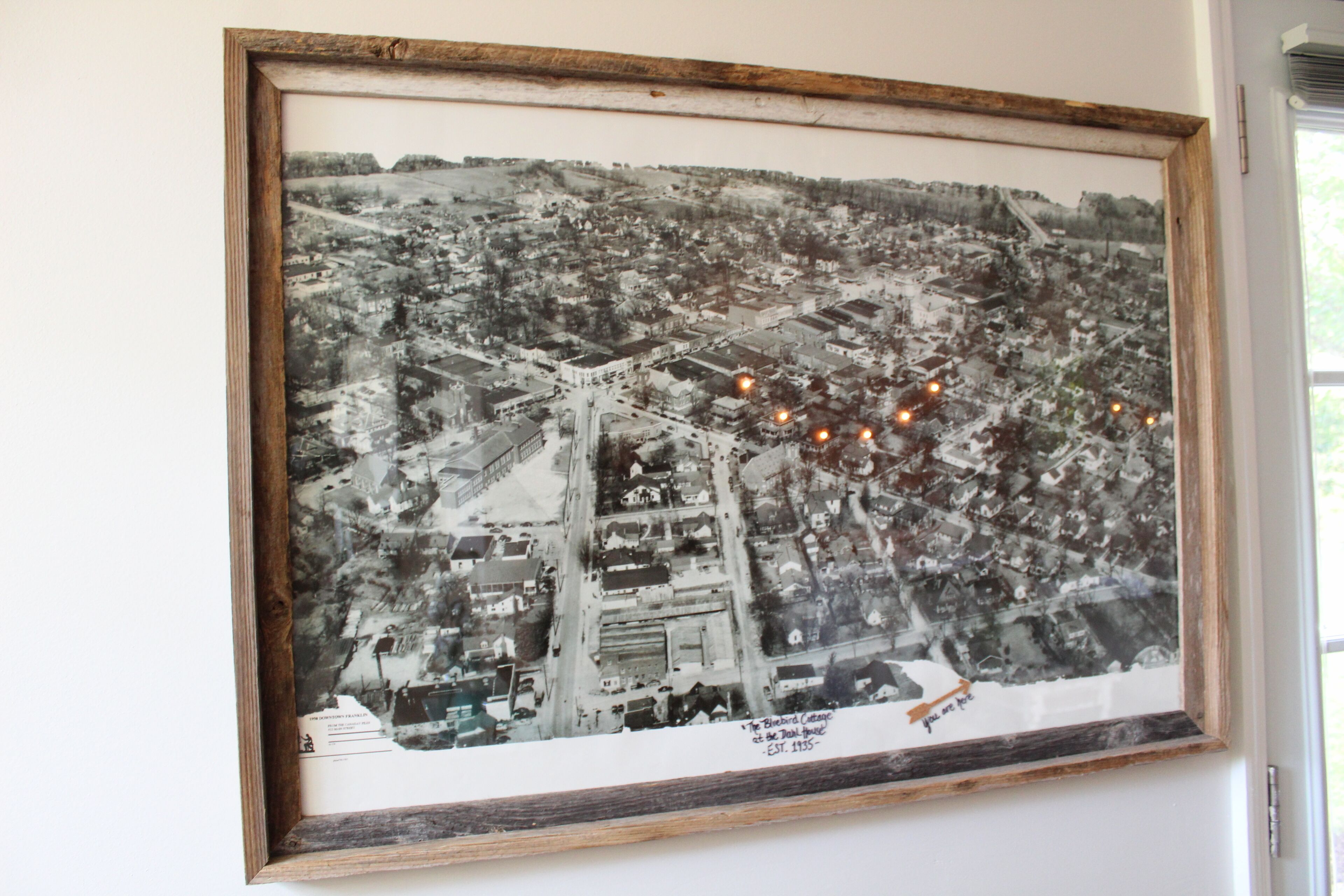 A framed black and white aerial photograph of a town with a prominent bridge and a sign indicating the establishment year.