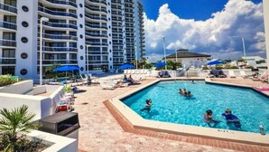 Uma área de piscina com espreguiçadeiras e guarda-sóis, cercada por um edifício alto com varandas.