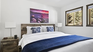 A bedroom with a bed, a nightstand, a lamp, a painting, and a window with a view of trees. - Outpost: Sleeping Indian E8 - Hot Tub Access! (Teton Village)