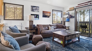 A living room with a brown leather sofa, a wooden coffee table, and a blue rug. There are two wall-mounted lamps and a window with a view of trees. - Outpost: Sleeping Indian E8 - Hot Tub Access! (Teton Village)