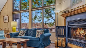 Un salón acogedor con un sofá azul, una mesa de centro de madera, una chimenea y ventanas amplias que dan a vistas de árboles.
