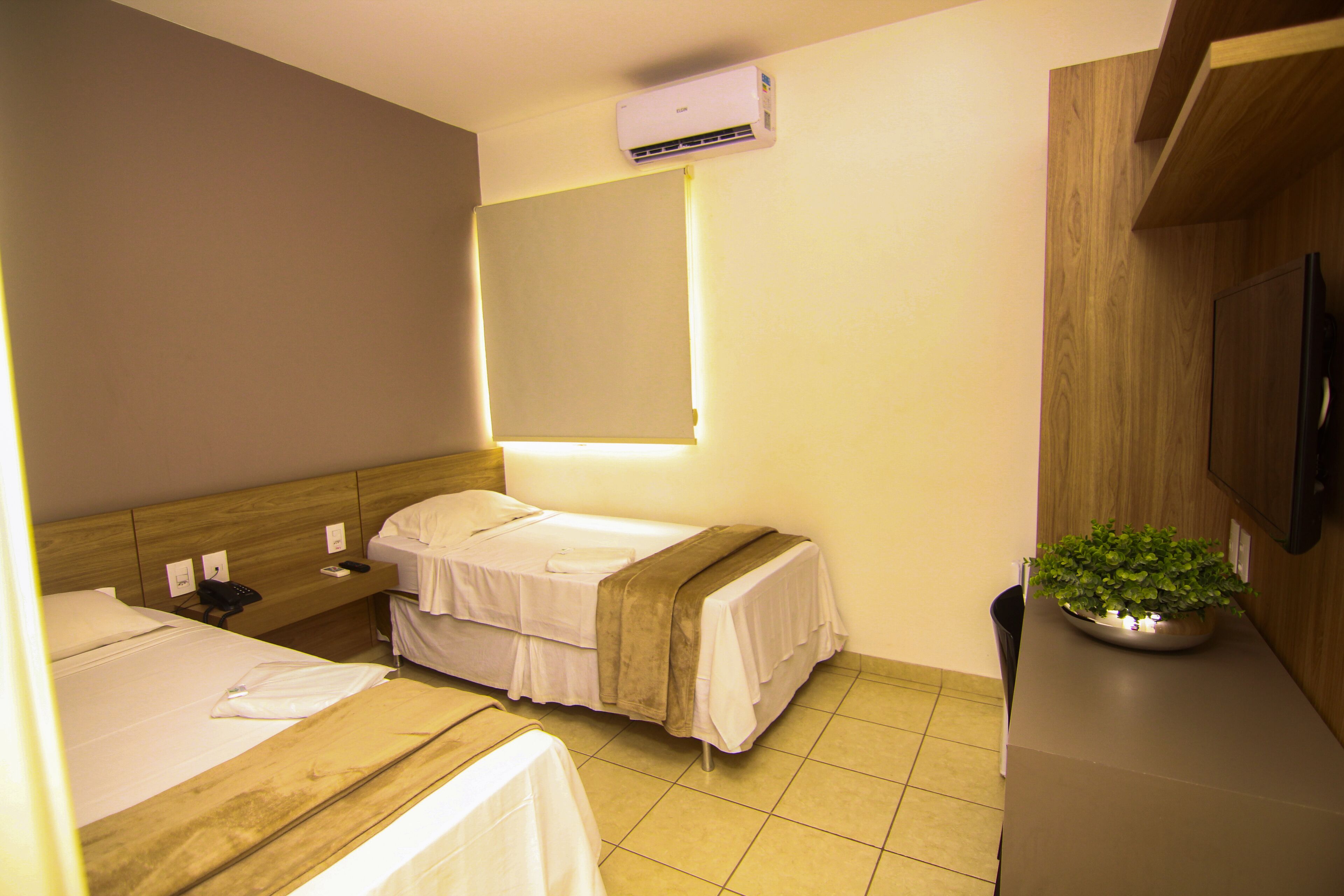 A hotel room with two beds, a bedside table with a lamp, a wall-mounted air conditioner, and a potted plant on the dresser.